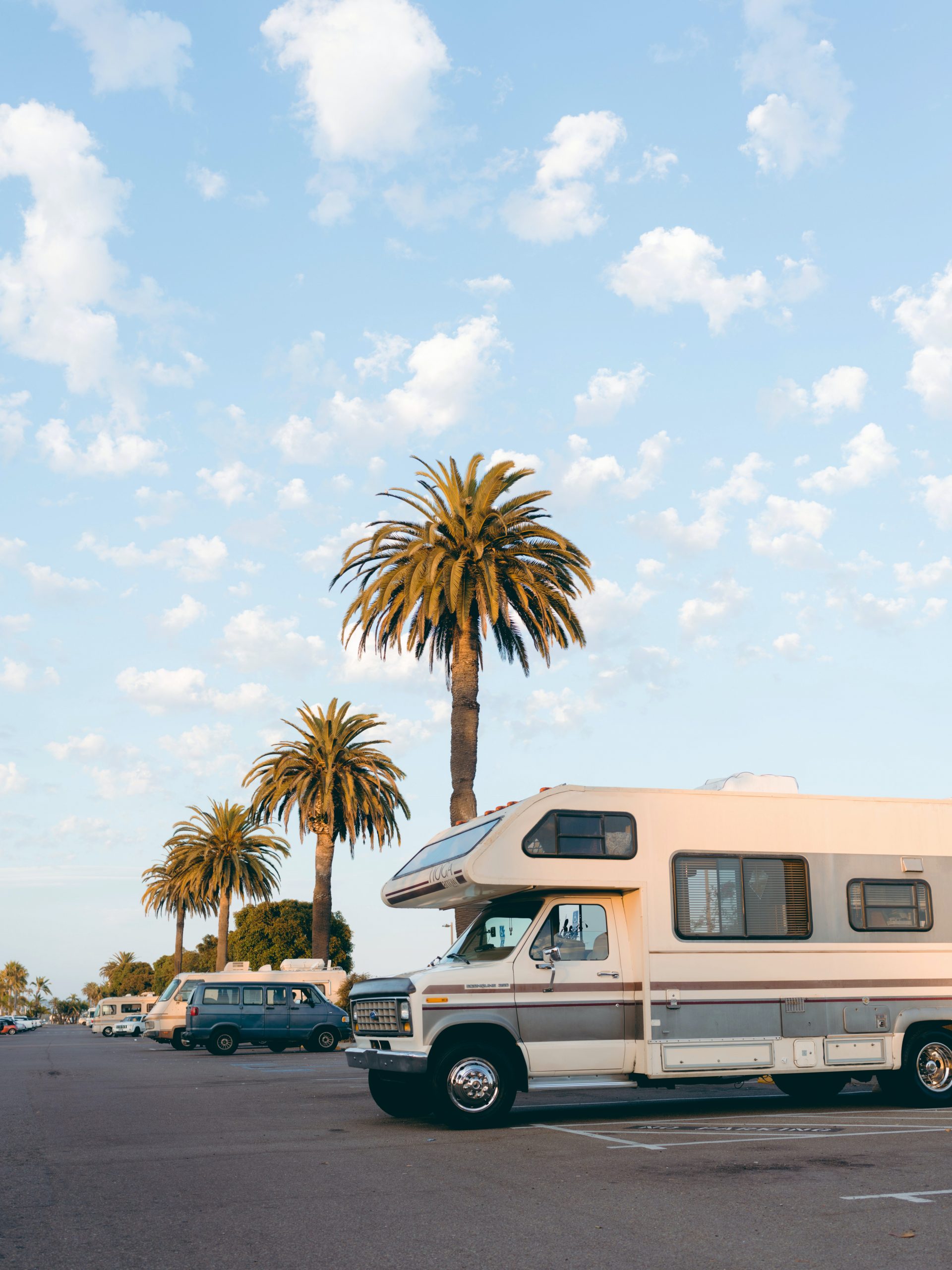 RV in parking lot with three palms trees angled down with light blue sky and white clouds.